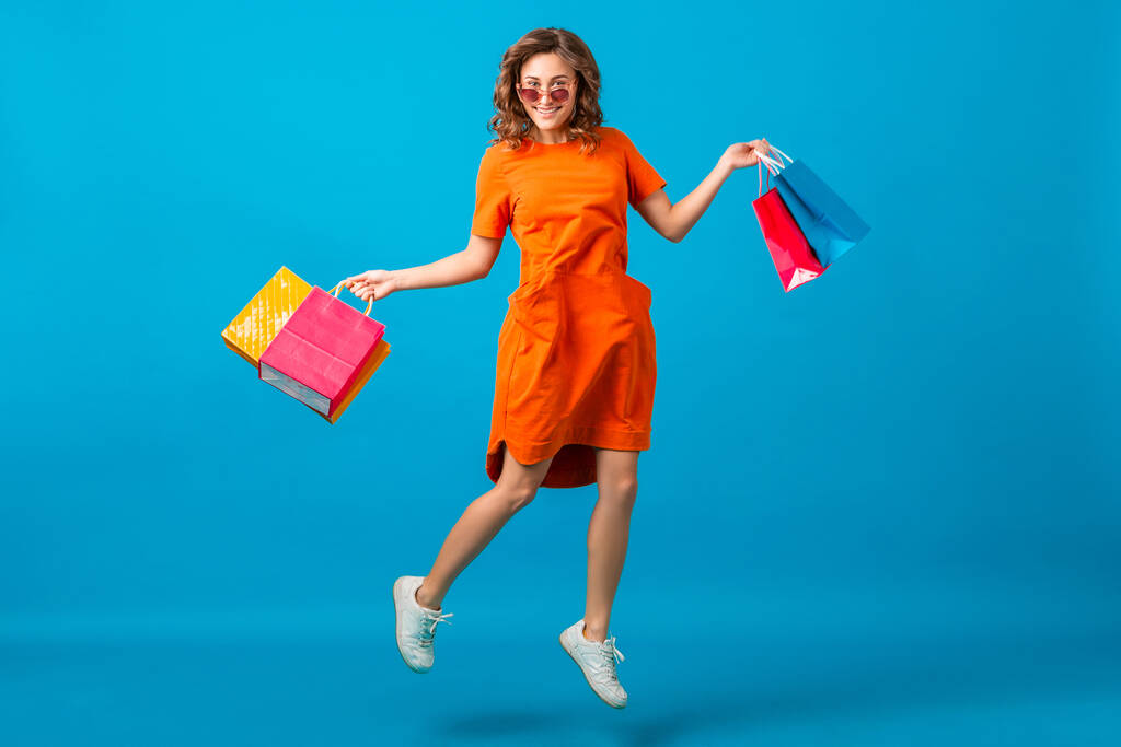 atrakcyjne szczęśliwy uśmiechnięty stylowy kobieta zakupoholiczka w pomarańczowy modny oversize sukienka skoki uruchomiony gospodarstwa torby na niebieskie studio tło odizolowane, kolorowe, sprzedaż podekscytowany, wiosna lato moda trend - Zdjęcie, obraz