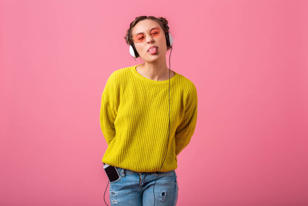vonzó boldog vicces nő hallgat zenét fülhallgató öltözött hipster színes stílusú ruhát elszigetelt rózsaszín stúdió háttér, visel sárga pulóver és napszemüveg, szórakozás - Fotó, kép