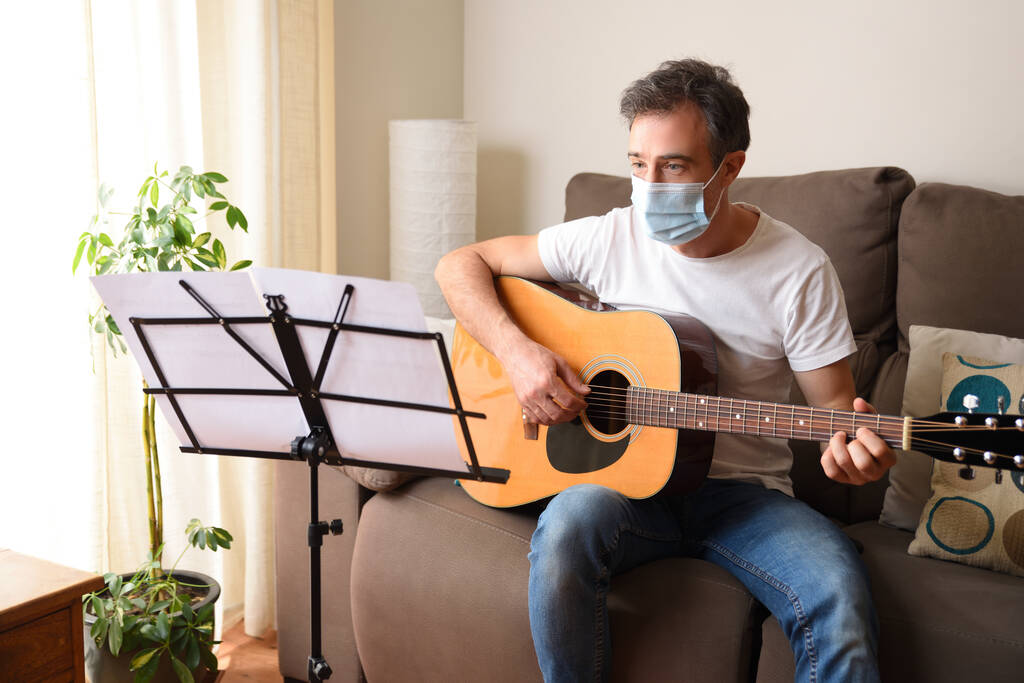 Felnőtt férfi maszkkal tanul akusztikus gitározni ül a kanapén a nappalijában részletesen - Fotó, kép
