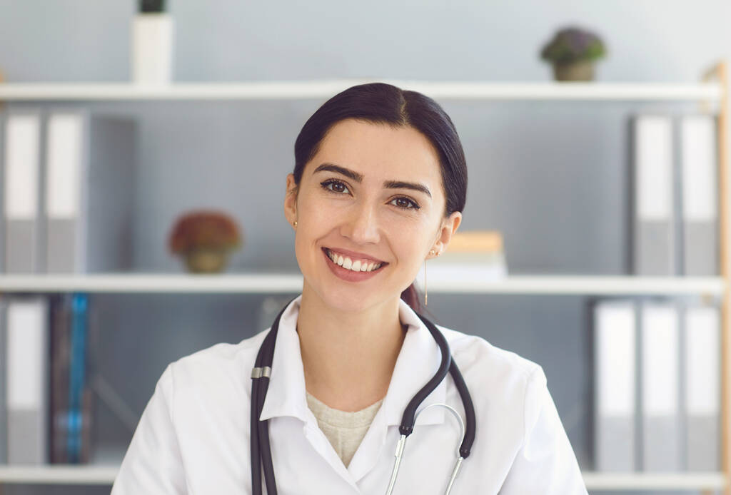 Портрет красивой молодой женщины-врача, смотрящей в камеру в медицинском кабинете, имеющей онлайн консультацию с пациентом
 - Фото, изображение