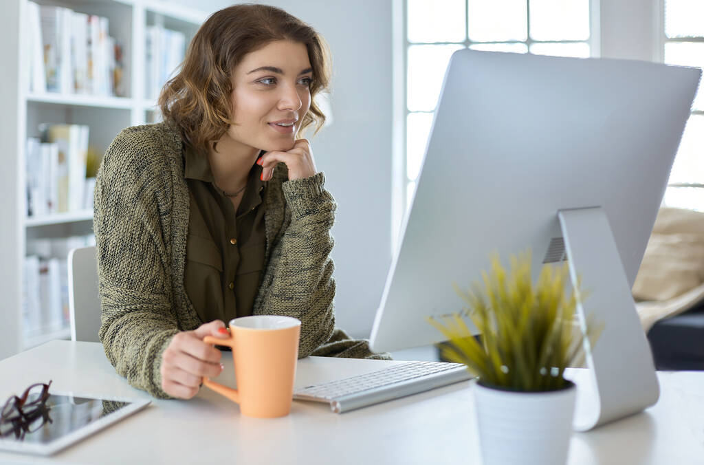 Çekici, gülümseyen bir kadın büro masasında oturuyor, elinde bir fincan kahve tutuyor, rahatlıyor ve başka tarafa bakıyor. - Fotoğraf, Görsel