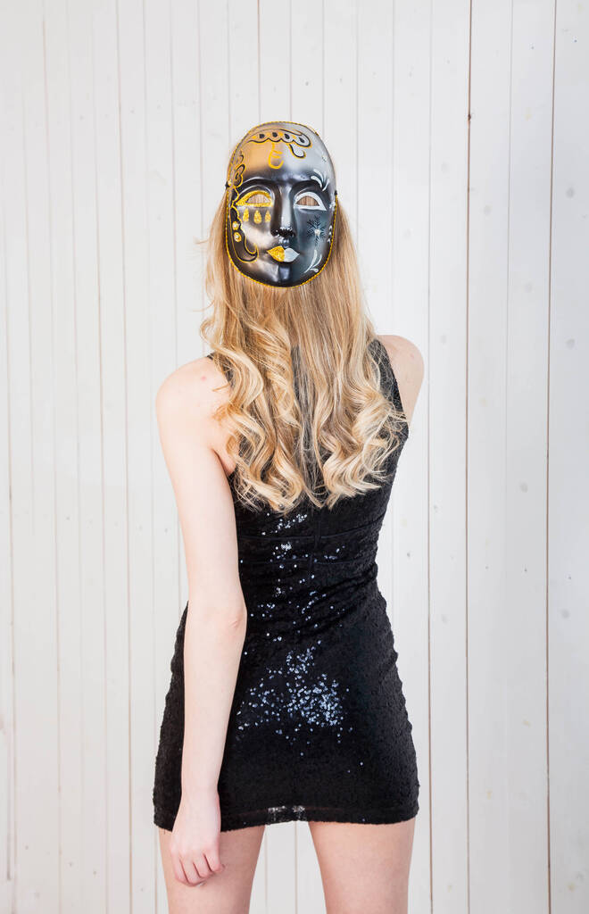 blond meisje in een zwart kort jurk staat met haar rug naar het podium dragen van een acteur masker op haar hoofd - Foto, afbeelding