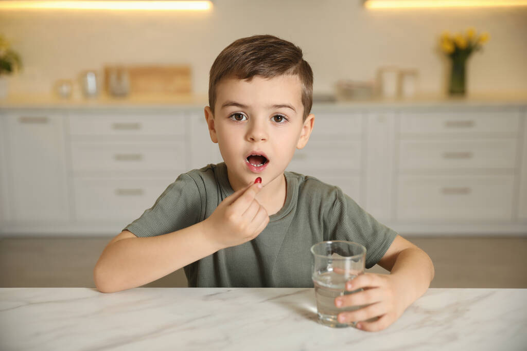 Menino com copo de água tomando cápsula de vitamina na cozinha
 - Foto, Imagem