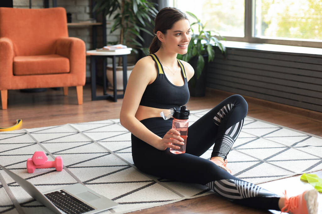 Fitness gyönyörű karcsú nő ül a padlón súlyzókkal és üveg vízzel laptoppal otthon a nappaliban. Maradj otthon! - Fotó, kép