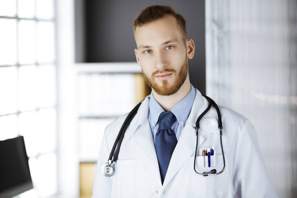 Medico con la barba rossa in piedi dritto in clinica vicino al suo posto di lavoro. Ritratto di medico. Medicina e concetto di assistenza sanitaria - Foto, immagini