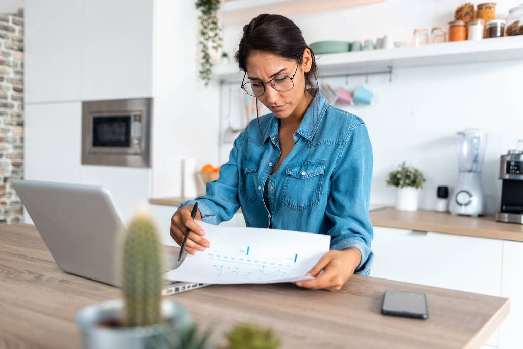 Prise de vue de jolie jeune femme examinant la paperasse et travaillant sur un ordinateur portable dans la cuisine à la maison. - Photo, image