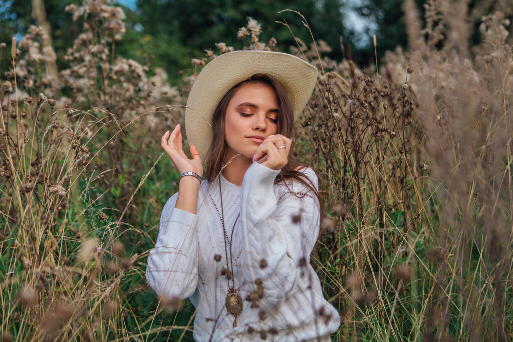 Hermosa chica adolescente morena vestida con un suéter blanco, vaqueros y sombrero de paja vaquero sentado en hierba de fresno marrón seco, sonriendo y riendo durante la puesta del sol
 - Foto, imagen