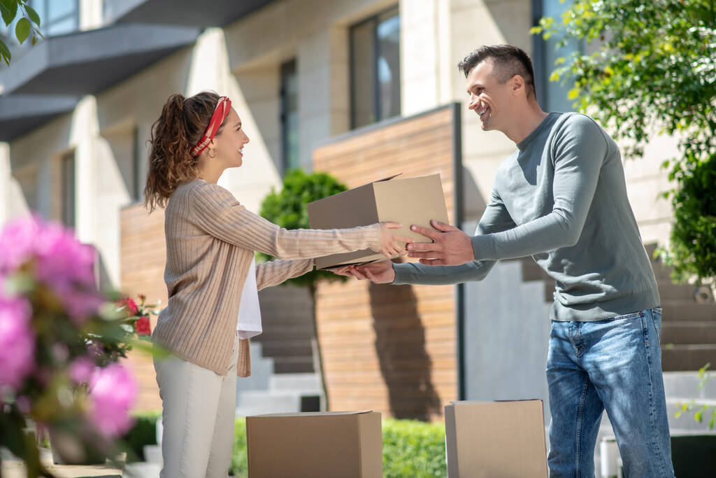 Γυναίκα με λευκό παντελόνι να δίνει χαρτόνι στον άντρα της ενώ μετακομίζει στο νέο σπίτι. - Φωτογραφία, εικόνα