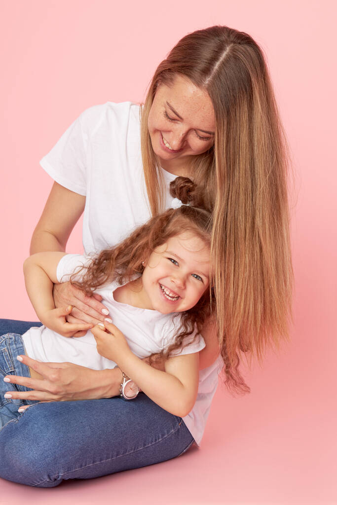 Семейные ценности. Мама и дочь в белых футболках и джинсах играют и обнимаются на розовом фоне. Заботиться о близких. Счастливое материнство
 - Фото, изображение
