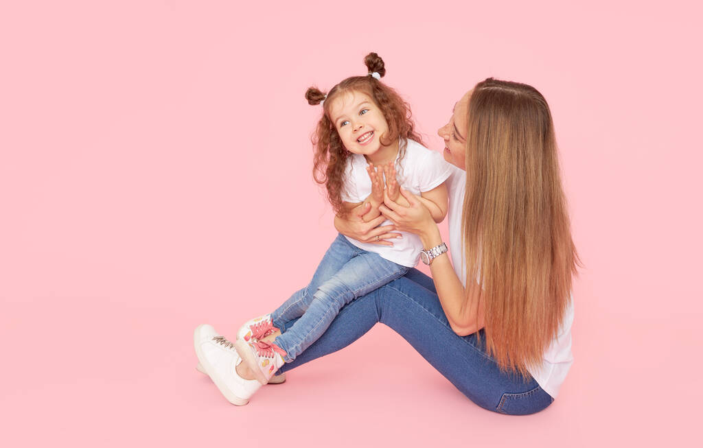 Gezinswaarden. Mam en dochter in witte t-shirts en jeans spelen en knuffelen op een roze achtergrond. Zorgen voor geliefden. Gelukkig moederschap - Foto, afbeelding