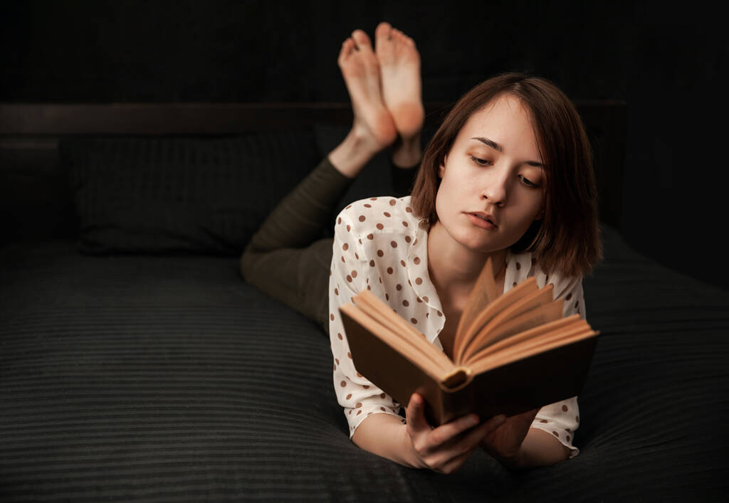 het meisje ligt met een boek in een zwart bed en leest zorgvuldig - Foto, afbeelding