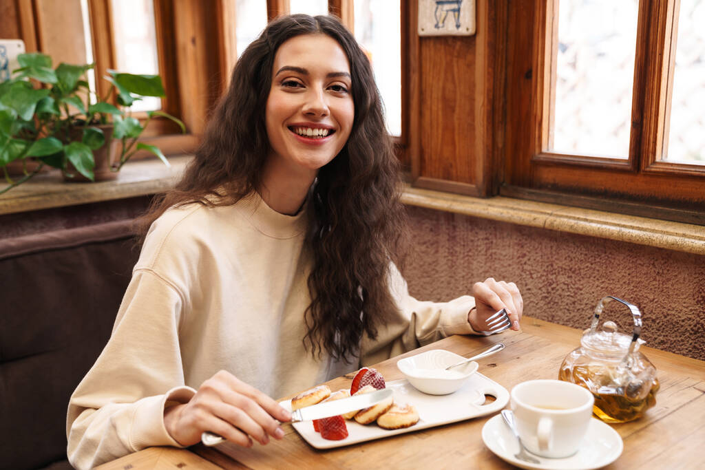 Imagen de la hermosa mujer joven feliz sonriendo y comiendo panqueques mientras está sentado en el acogedor café en el interior
 - Foto, imagen