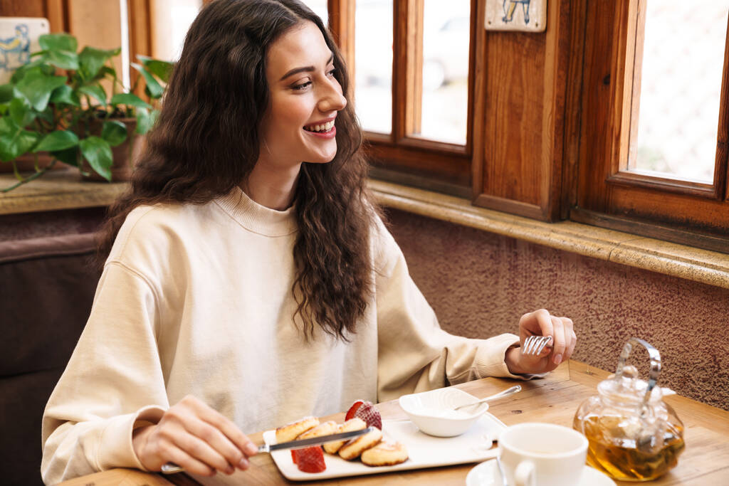 Изображение счастливой красивой молодой женщины, улыбающейся и поедающей блины, сидя в уютном кафе в помещении
 - Фото, изображение