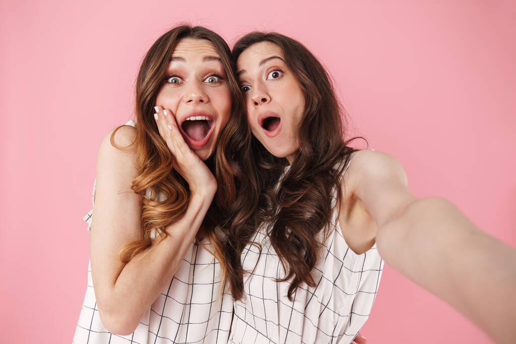Imagen de mujeres guapas emocionadas expresando sorpresa mientras toman fotos de selfies aisladas sobre fondo rosa
 - Foto, imagen