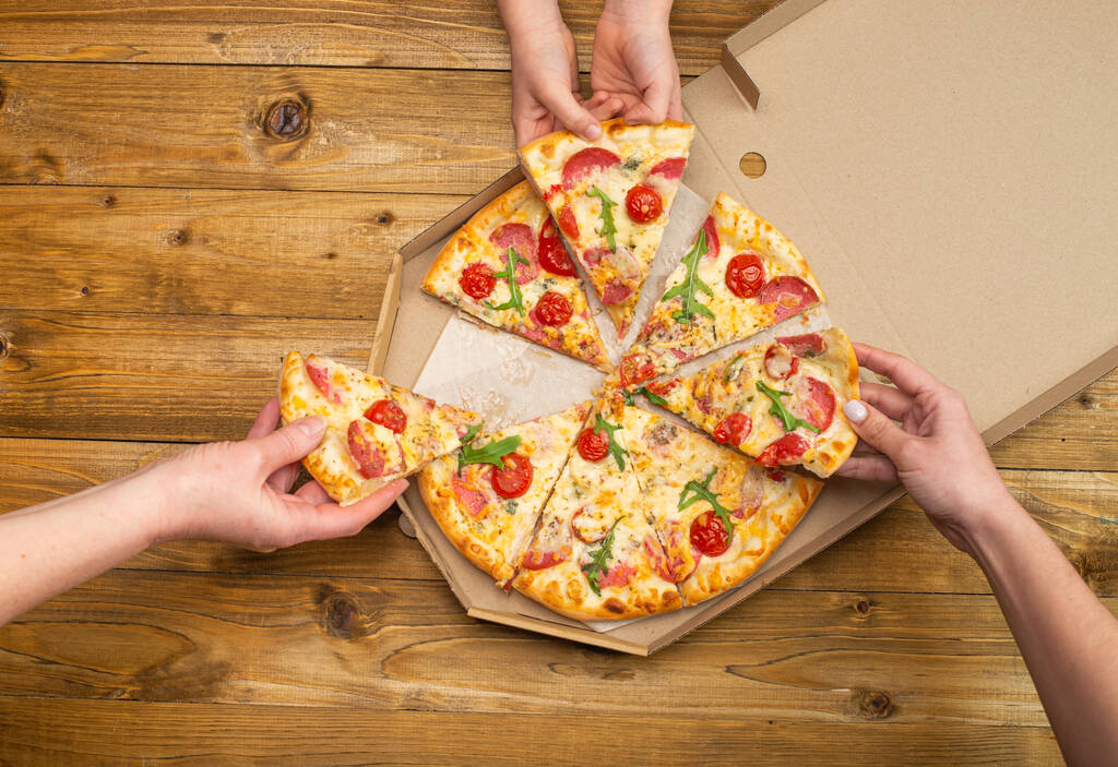 bijgesneden shot van mensen met plakjes pizza met tomaten, salami en kaas op houten tafel - Foto, afbeelding