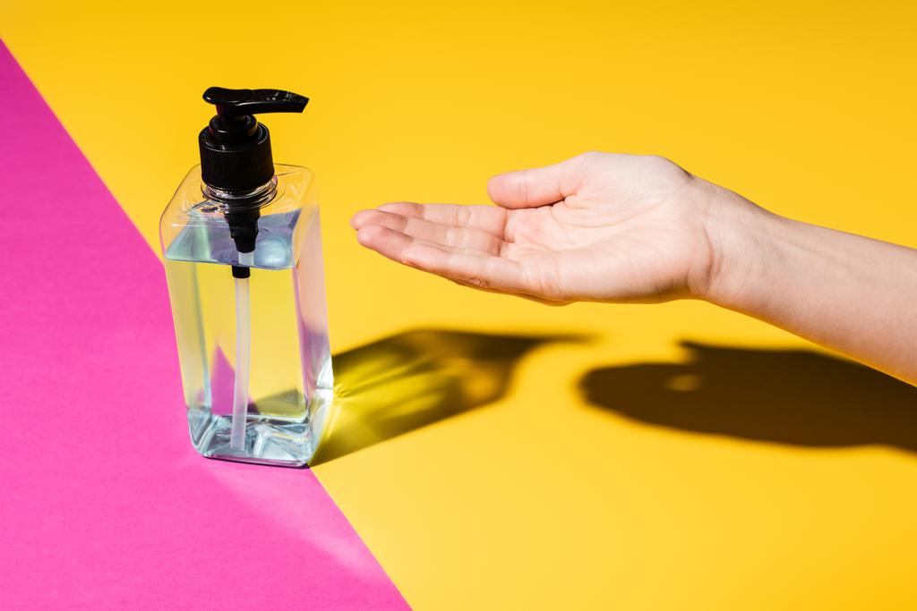 Teilansicht der weiblichen Hand in der Nähe der Flasche mit Desinfektionsmittel auf rosa und gelb - Foto, Bild