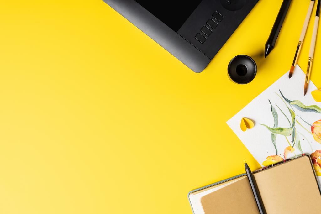 bovenaanzicht van penselen in de buurt van schilderen, tekentablet, notebook en stylus op geel  - Foto, afbeelding