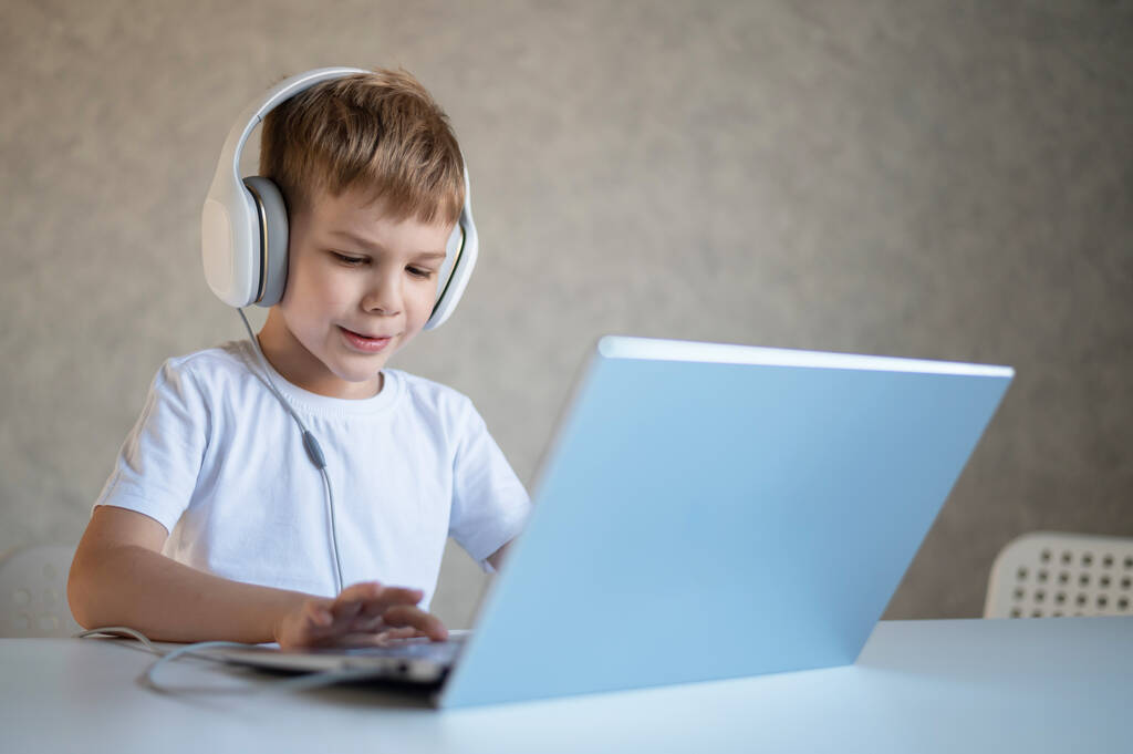 Χαριτωμένο ευρωπαϊκό αγόρι ακούει μουσική σε ακουστικά και τύπους σε ένα φορητό υπολογιστή. Το παιδί διαβάζει στον υπολογιστή. Ένας νεαρός μαθητής λαμβάνει εκπαίδευση εξ αποστάσεως. - Φωτογραφία, εικόνα