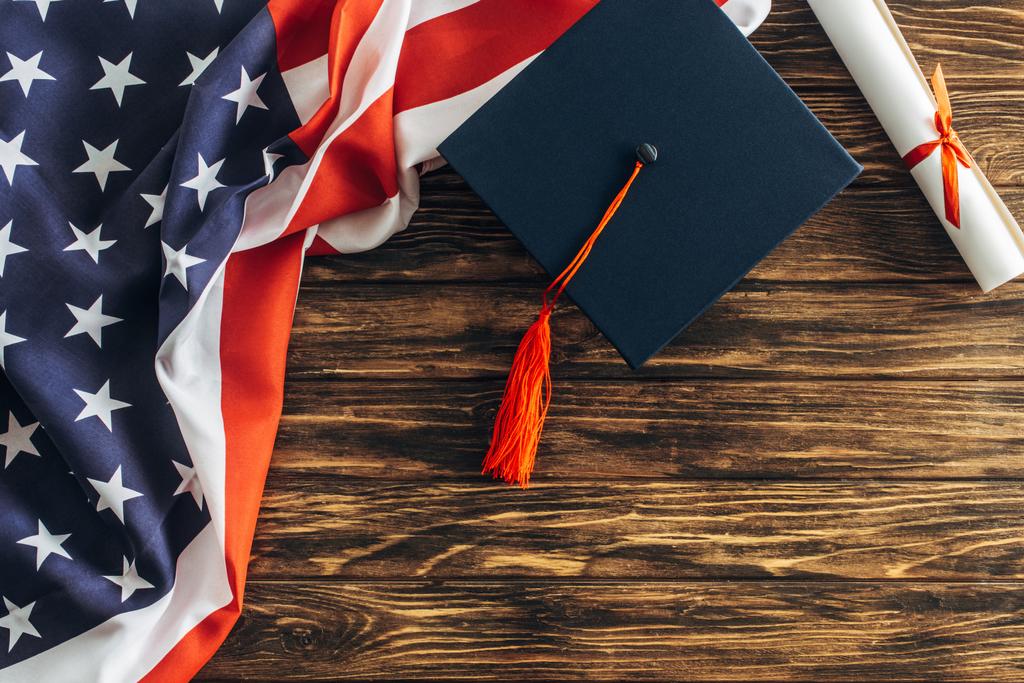 vista superior do diploma e tampa de graduação perto da bandeira americana com estrelas e listras na superfície de madeira
  - Foto, Imagem