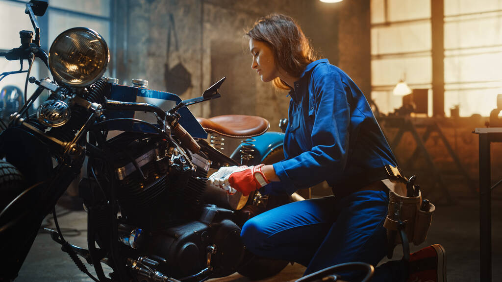 Junge schöne Mechanikerin arbeitet an einem Custom-Motorrad in der Garage. Talentierte Mädchen tragen einen blauen Overall. Sie benutzt eine Ratsche, um Mutternschrauben festzuziehen. Kreativ-authentische Werkstatt. - Foto, Bild