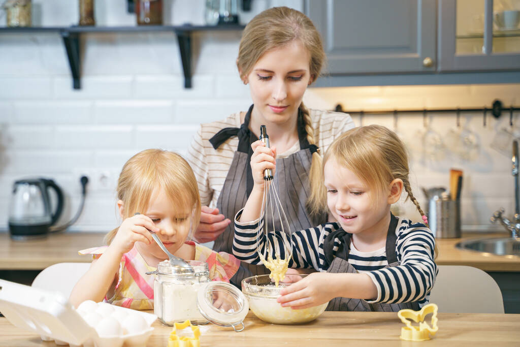 Ευτυχισμένη οικογένεια, νεαρή μητέρα και δύο κόρες που φτιάχνουν ζύμη για τηγανίτες ή μπισκότα μαζί στην κουζίνα.. - Φωτογραφία, εικόνα