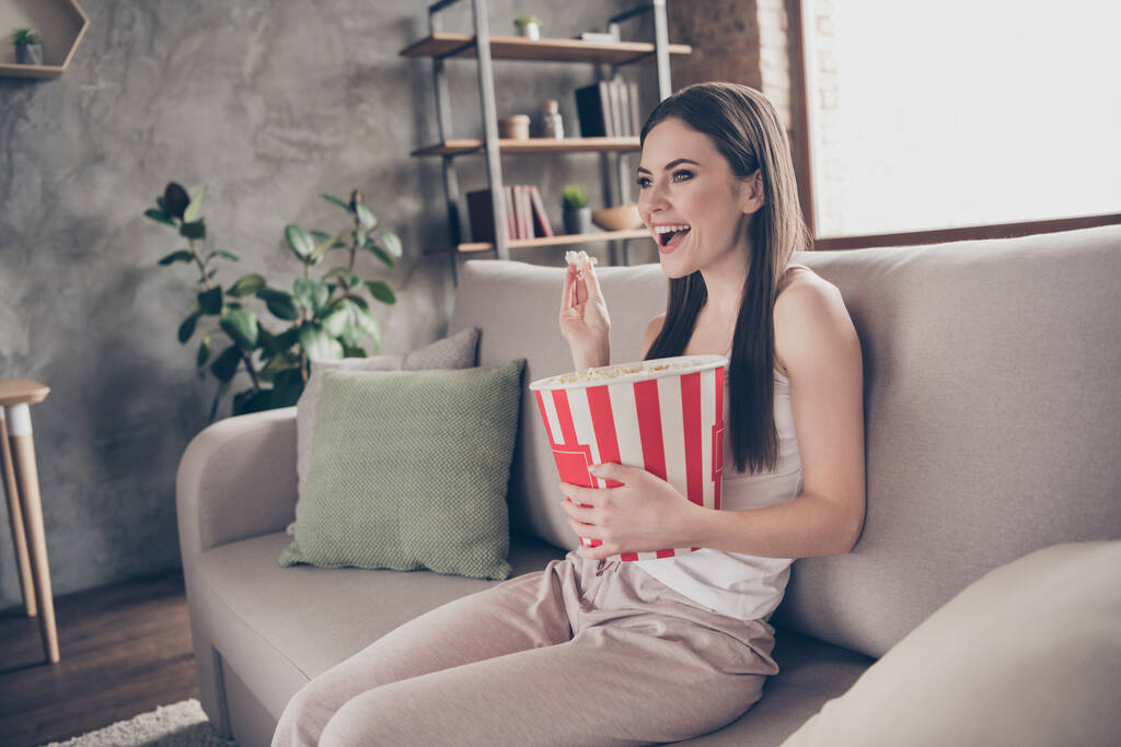 Profil côté photo de joyeux fille franche assis divan tenir grande boîte manger pop maïs regarder intéressant film série rester à la maison dans l'appartement maison à l'intérieur - Photo, image
