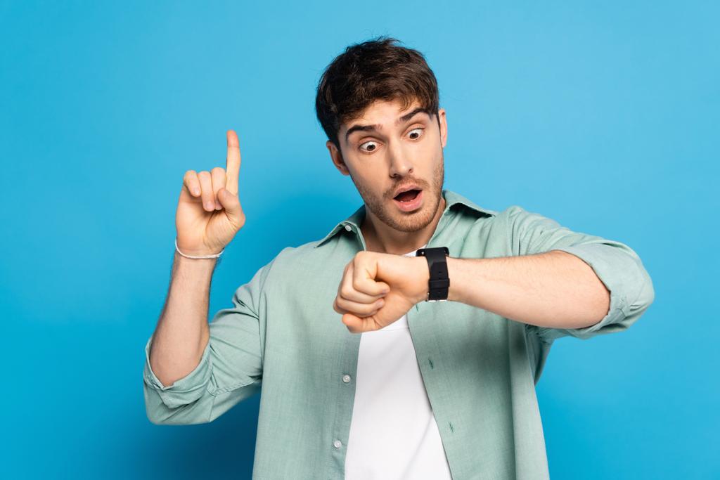 zszokowany młody człowiek pokazując gest idei patrząc na zegarek na niebiesko - Zdjęcie, obraz