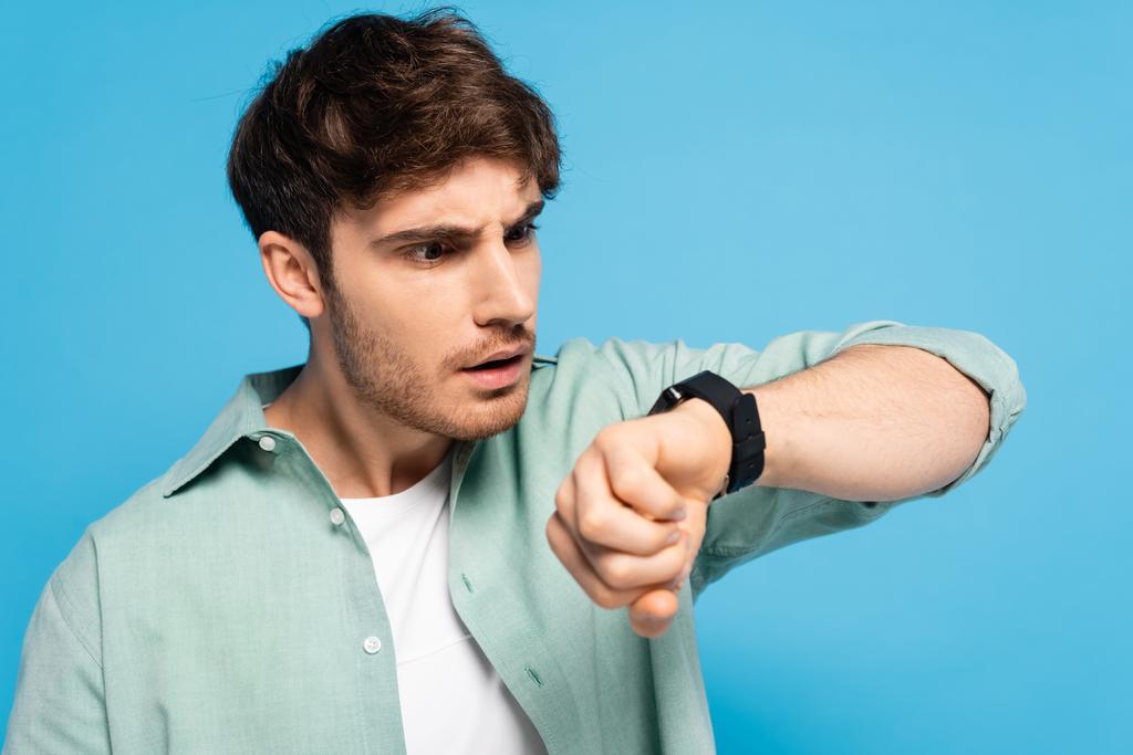 scioccato giovane uomo guardando orologio da polso isolato su blu - Foto, immagini