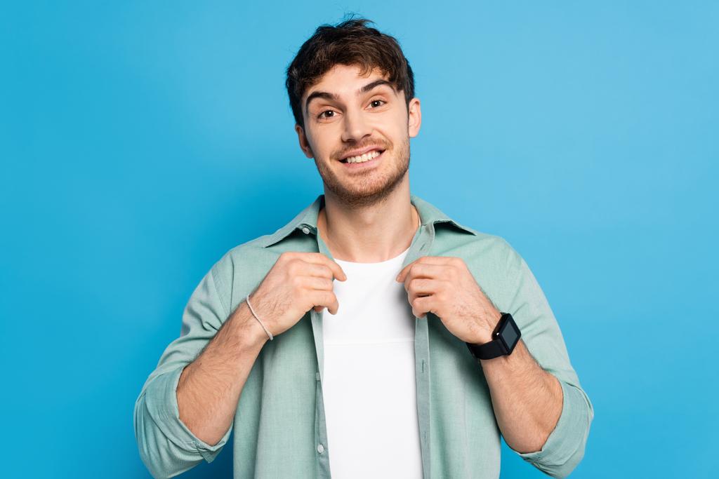 guapo joven tocando camisa mientras sonríe a la cámara en azul
 - Foto, Imagen