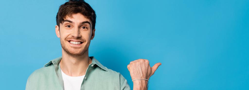 poziomy obraz szczęśliwego młodego człowieka wskazującego kciukiem na niebieski - Zdjęcie, obraz