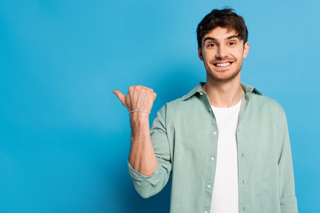 šťastný mladý muž ukazuje palcem při pohledu na kameru na modré - Fotografie, Obrázek