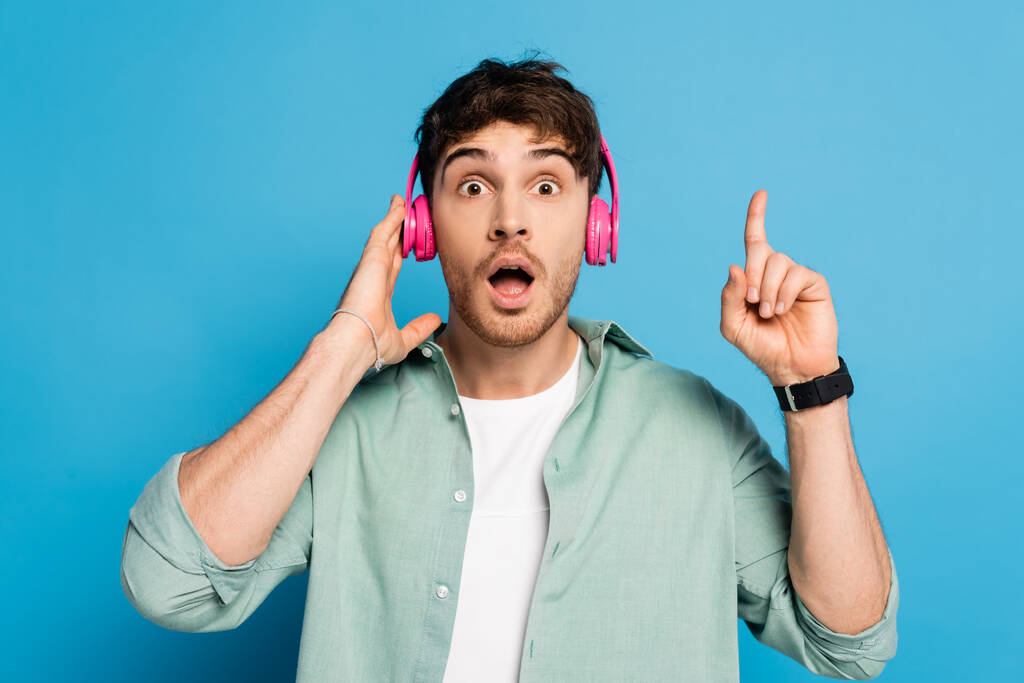 σοκαρισμένος νεαρός άνδρας αγγίζει ασύρματα ακουστικά και δείχνει χειρονομία ιδέα στο μπλε - Φωτογραφία, εικόνα