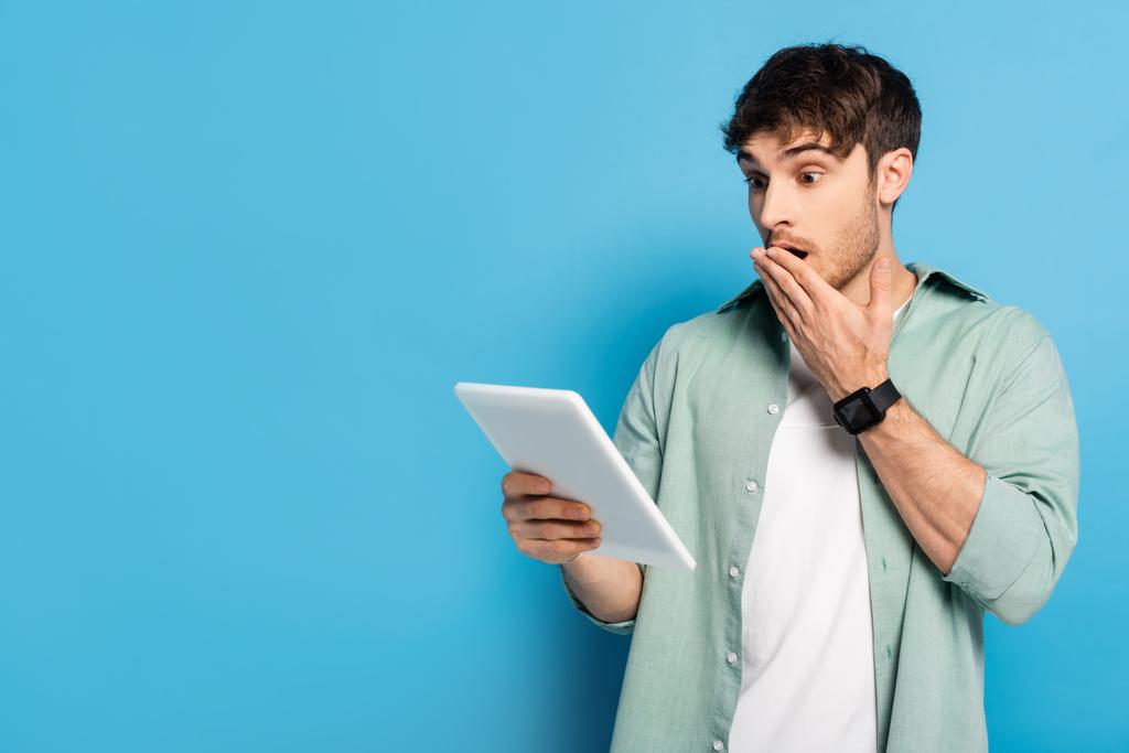 shocked young man covering mouth with hand while using digital tablet on blue - Photo, Image