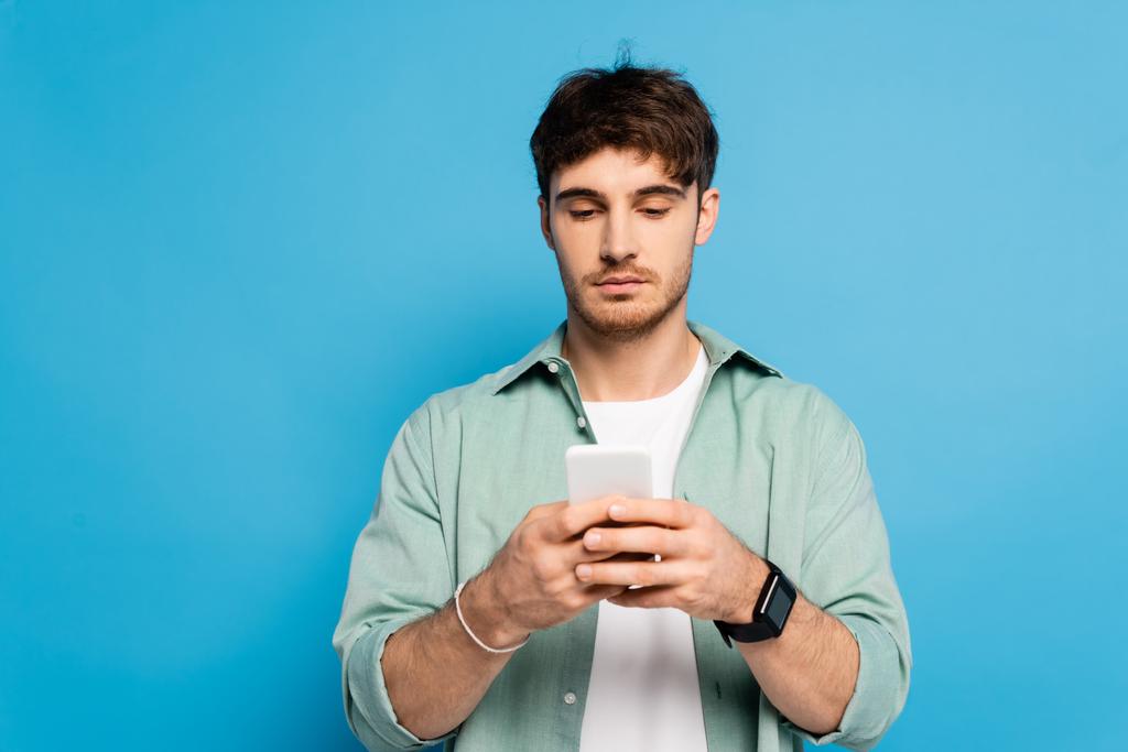 σοβαρός νεαρός συνομιλεί σε smartphone απομονωμένο σε μπλε - Φωτογραφία, εικόνα