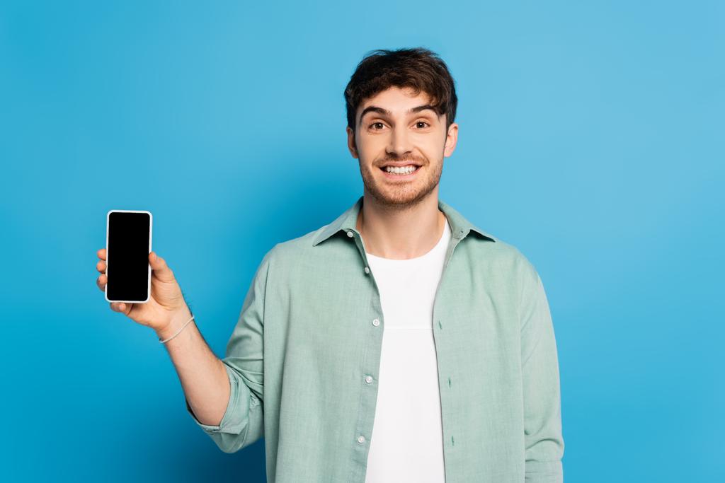 Glücklicher junger Mann zeigt Smartphone mit leerem Bildschirm auf blau - Foto, Bild