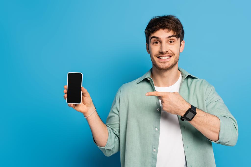 joven feliz apuntando al teléfono inteligente con pantalla en blanco en azul
 - Foto, imagen