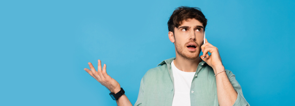 horizontální obraz zmateného mladého muže, který ukazuje gesto pokrčení ramen, zatímco mluví na smartphonu izolovaném na modré - Fotografie, Obrázek