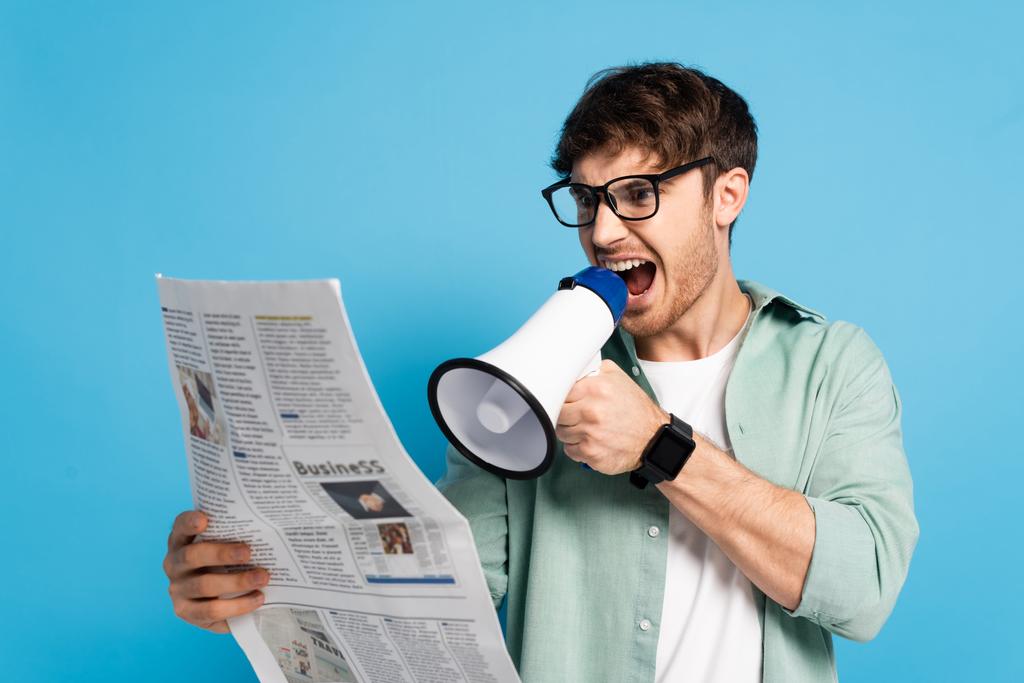 rozzlobený mladý muž křičí v megafonu při čtení novin na modré - Fotografie, Obrázek