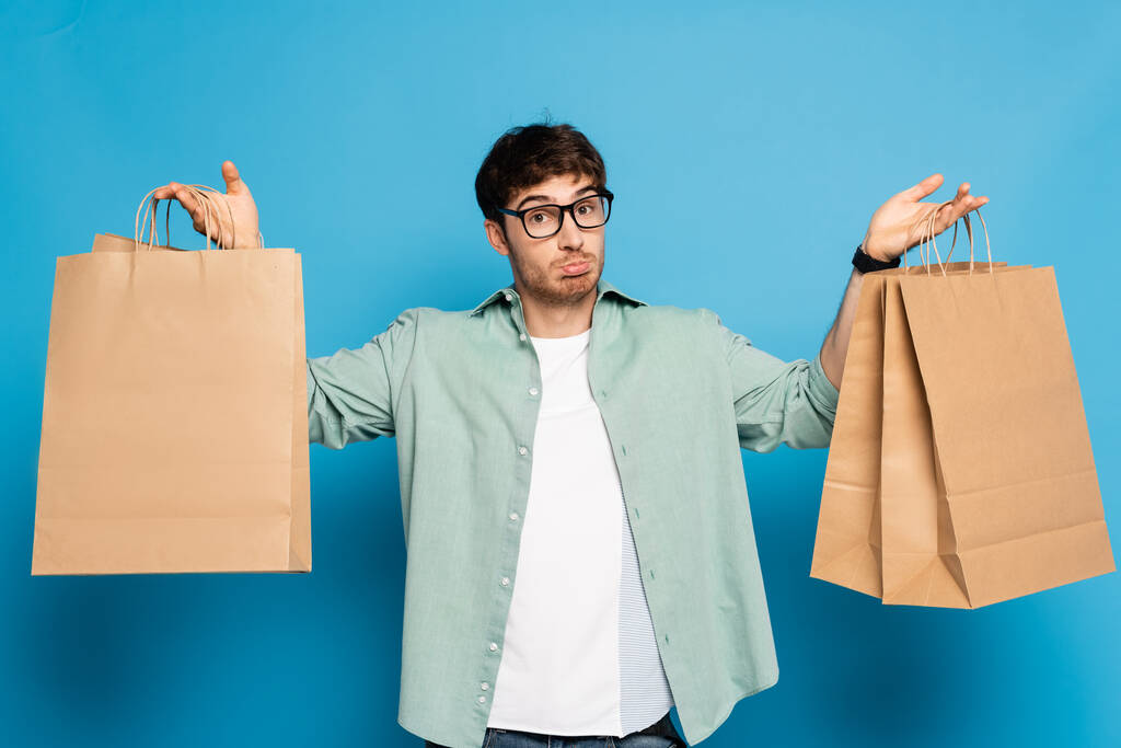 Alışveriş torbaları tutan kafası karışmış genç bir adam mavi kameraya bakarken - Fotoğraf, Görsel