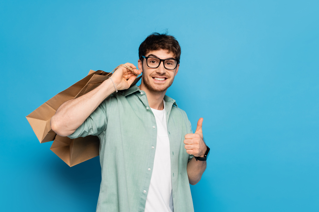 szczęśliwy młody człowiek pokazując kciuk w górę podczas noszenia torby na zakupy na niebieski - Zdjęcie, obraz
