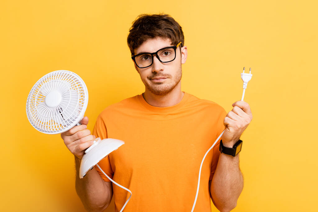 разочарованный молодой человек держит электрический вентилятор, глядя на камеру на желтом
 - Фото, изображение