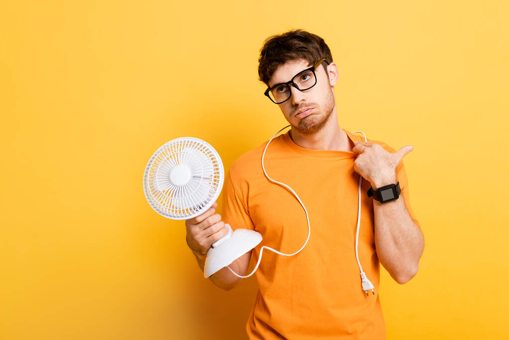 elégedetlen férfi gazdaság elektromos ventilátor, miközben szenved a hő sárga - Fotó, kép