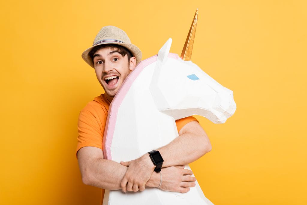 excited man in summer hat hugging decorative unicorn on yellow  - Photo, Image