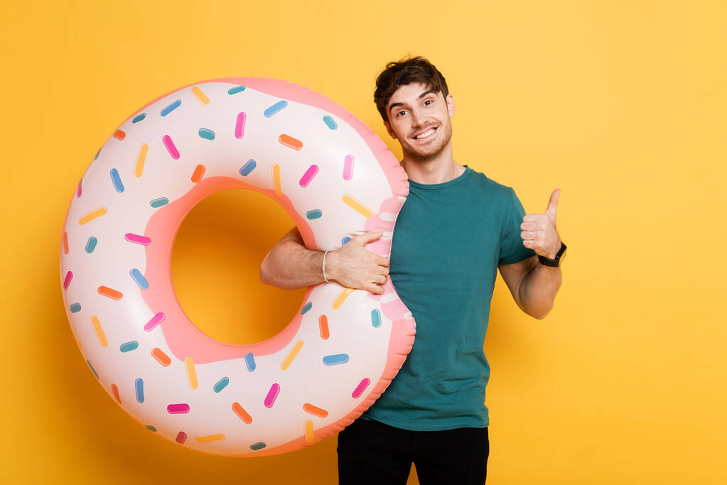 fröhlicher Mann zeigt Daumen hoch und steht mit aufblasbarem Donut auf gelb  - Foto, Bild