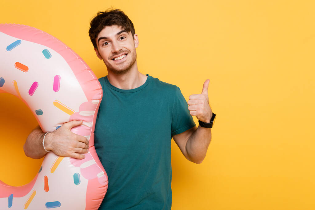 Lächelnder Mann zeigt Daumen nach oben, während er mit aufblasbarem Donut auf gelb steht  - Foto, Bild
