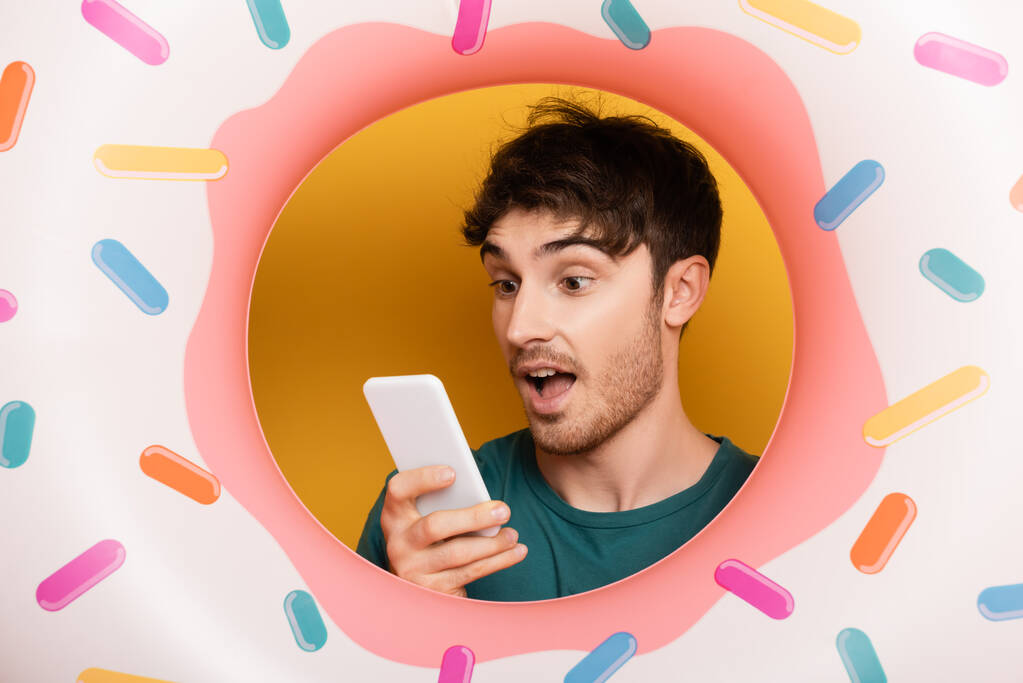 surpris jeune homme avec beignet gonflable en utilisant un smartphone sur jaune - Photo, image
