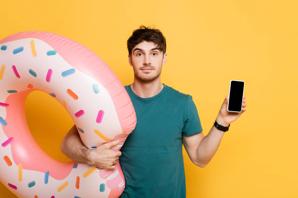 jovem com donut inflável segurando smartphone com tela em branco no amarelo
 - Foto, Imagem