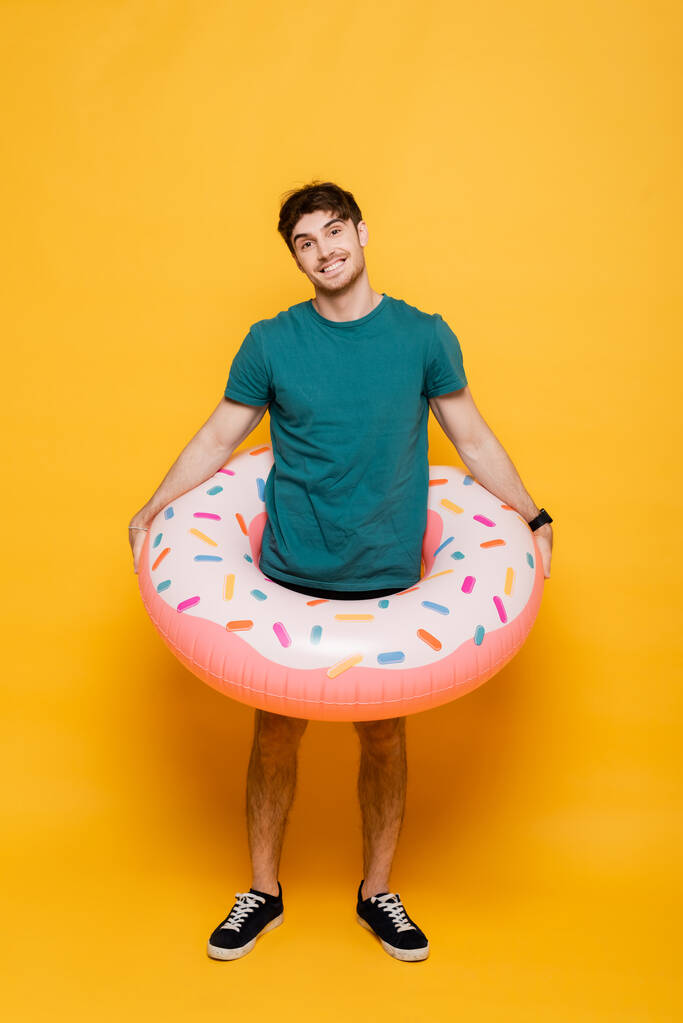 glimlachende man staand met opblaasbare donut op geel  - Foto, afbeelding