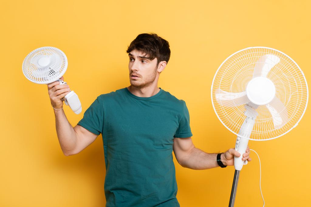 giovane uomo frustrato in possesso di due ventilatori elettrici su giallo    - Foto, immagini