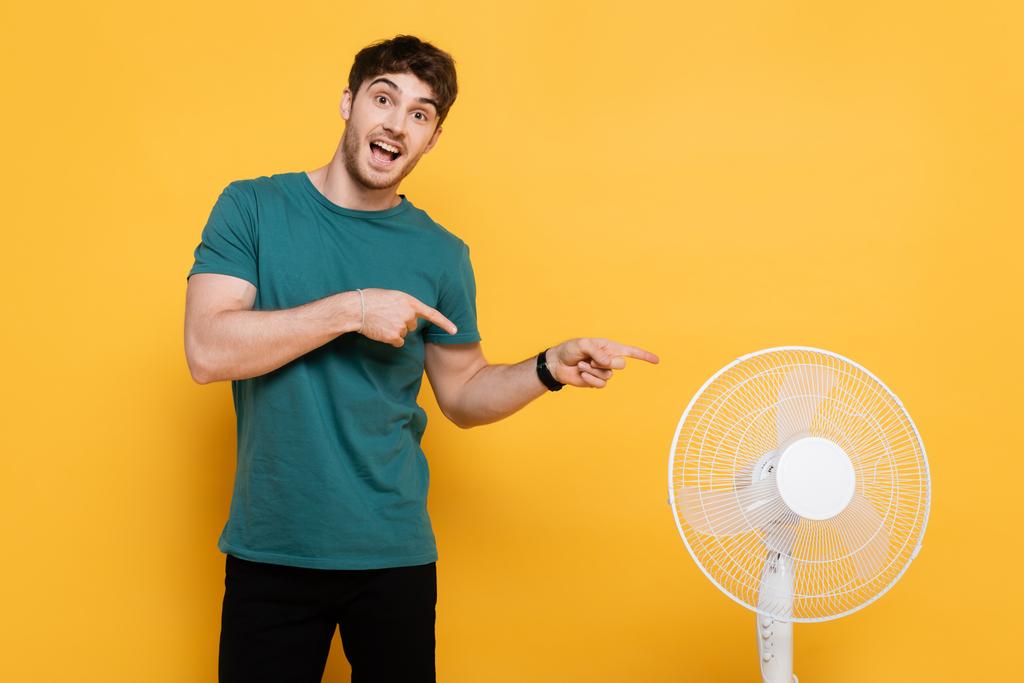 cheerful young man pointing with fingers at electric fan on yellow     - Photo, Image